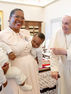 Pope Francis, dressed in white papal vestments and holding a cane, smiles and points toward a joyful Anita Annet Among wearing glasses and a light-colored dress with a large bow. She is holding her twins dressed in white outfits; one baby looks directly at the camera while the other rests against her. The scene takes place in an elegant, ornate room with religious artwork, wood paneling, and patterned rugs.