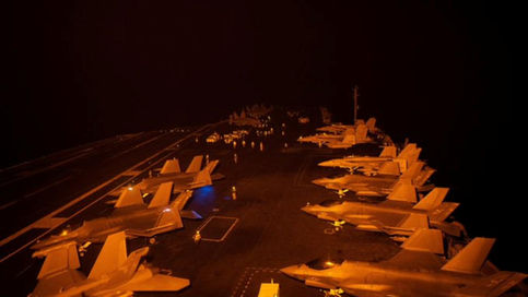 Nighttime view of multiple F-35 fighter jets parked on the illuminated flight deck of a U.S. aircraft carrier, with crew members and deck equipment visible under orange lighting against a dark sky.