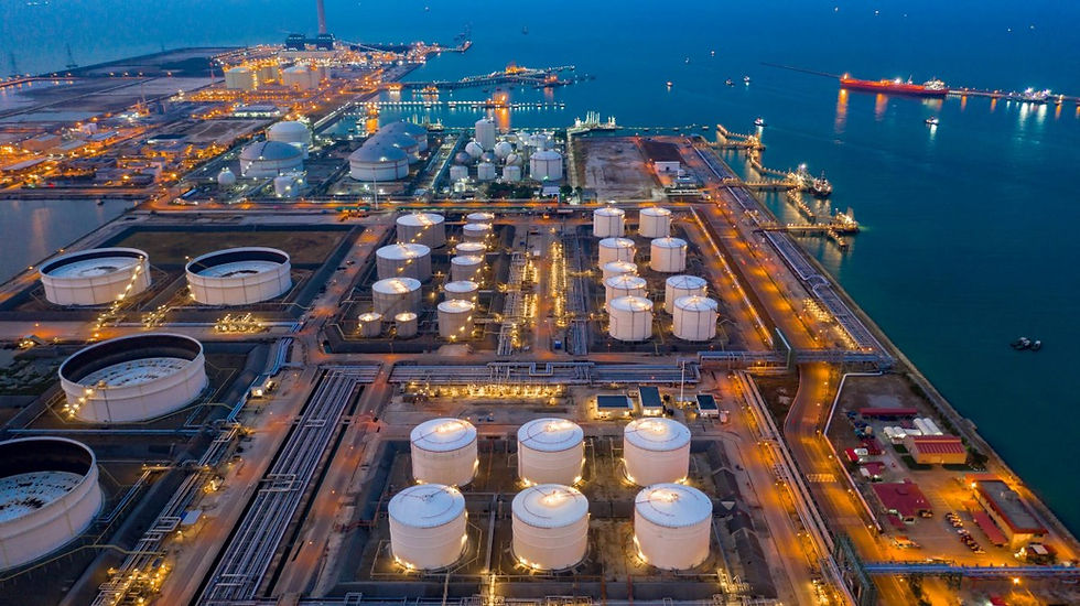 High-angle aerial view at dusk of a sprawling coastal petrochemical refinery and oil storage facility next to a deepwater shipping harbor. Rows of illuminated large white circular oil storage tanks are connected by a network of pipelines and industrial roads, with cargo ships anchored in the calm ocean water in the background under a dim evening sky.