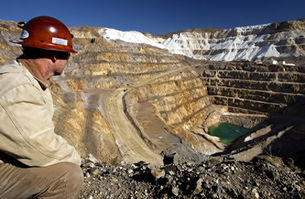 A miner in red hard hat crouching at the edge of a massive open-pit mine with white ash deposits along the ridges and terraces, highlighting the scale of rare earth and critical mineral mining operations.