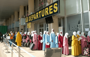 Uganda women in colorful robes, line up at Entebbe airport departures area. A large yellow-and-black "DEPARTURES" sign is visible above them. A quiet testament to Uganda’s reliance on labour export as a pathway to opportunity abroad.
