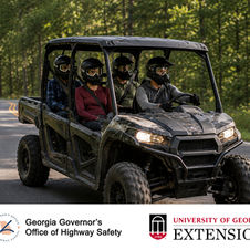 Photo of a black UTV carrying four riders on a paved road through a wooded area. All four riders are wearing helmets, and the driver and passengers are secured with seat belts. The bottom of the graphic displays the logos of the Georgia Governor’s Office of Highway Safety and University of Georgia Extension.