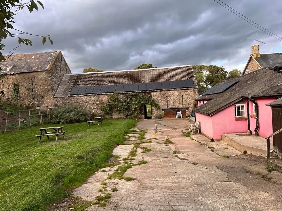Outdoor photography of a rustic stone barn with solar panels on its roof, adjacent to a bright pink building. The scene is set on a lush, overcast day with a worn paved path leading towards the structures.