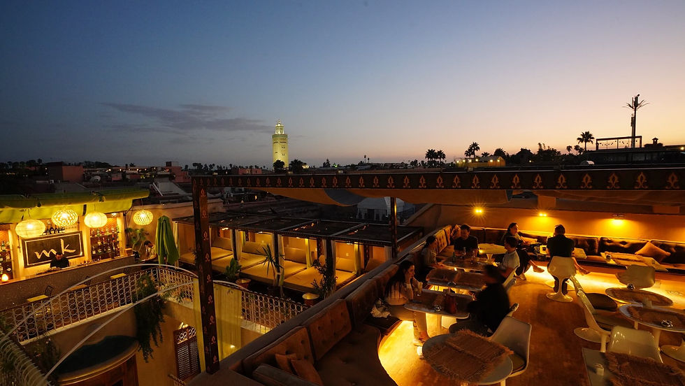 Dakterraslounge bij zonsondergang met dinerende mensen, warme verlichting en uitzicht op de stad met een verlichte toren. Gezellige, uitnodigende sfeer.