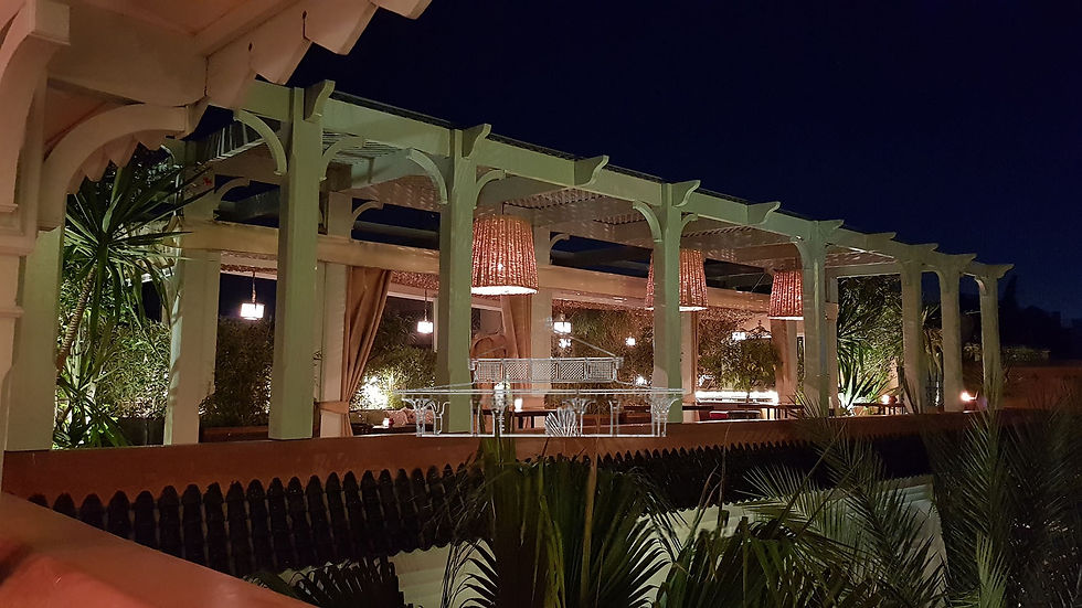Avondterras met verlichte pergola, gevlochten hanglampen, groen en decoratieve pilaren. Warme sfeerverlichting zorgt voor een gezellige sfeer.
