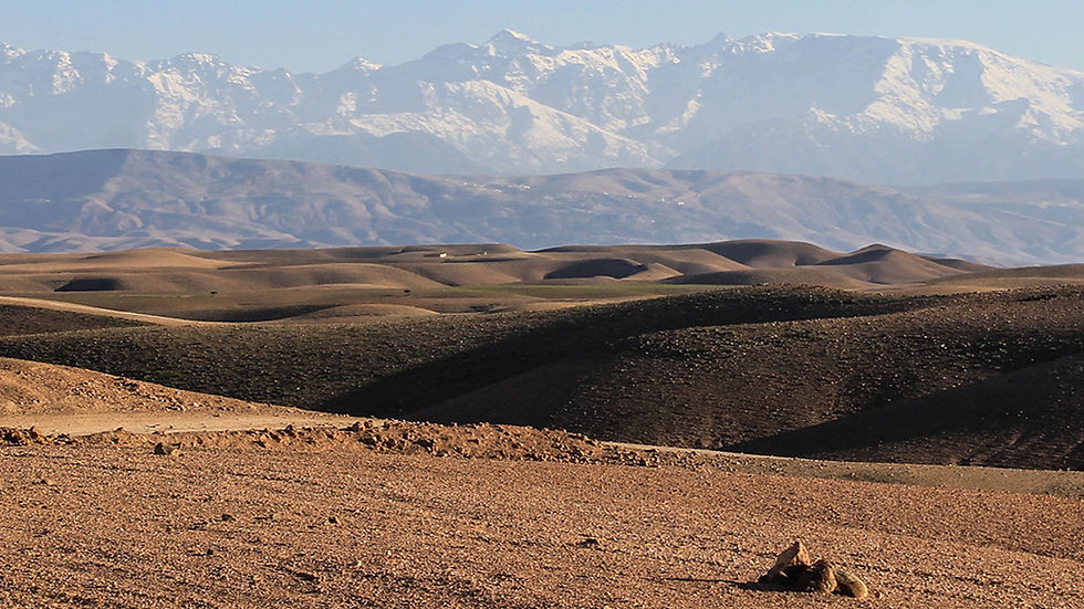 Arid rolling brown hills in the foreground, dark ridges, and snowcapped mountains under a hazy blue sky.