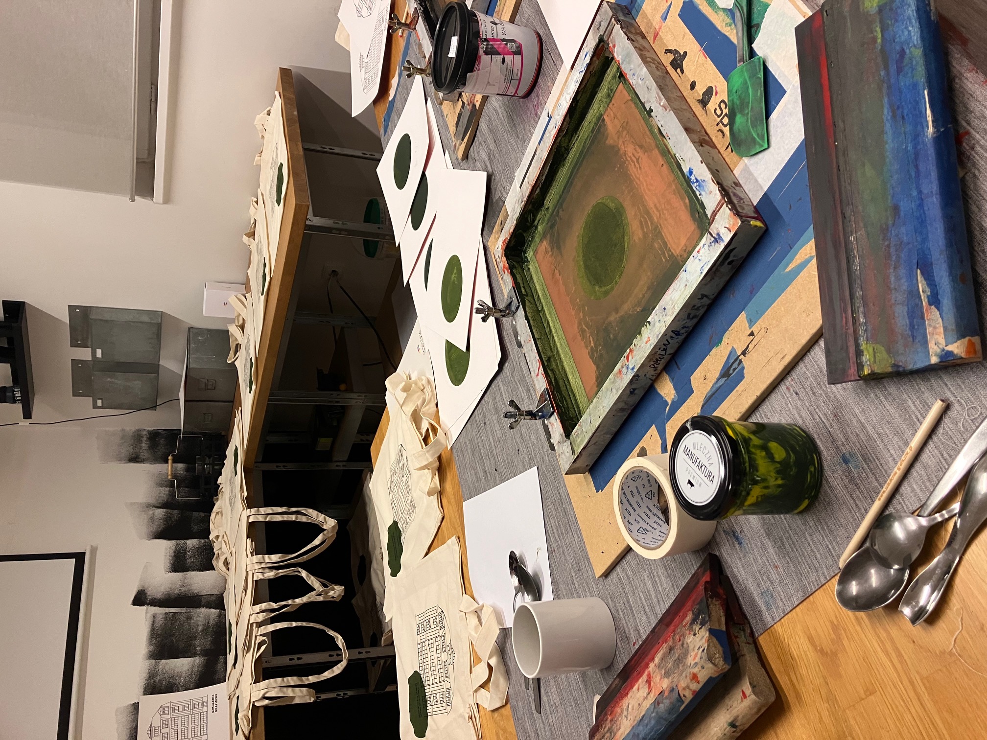 Pracownia sitodruku: sita, rakle, farby oraz torby i odbitki przygotowane na stołach.