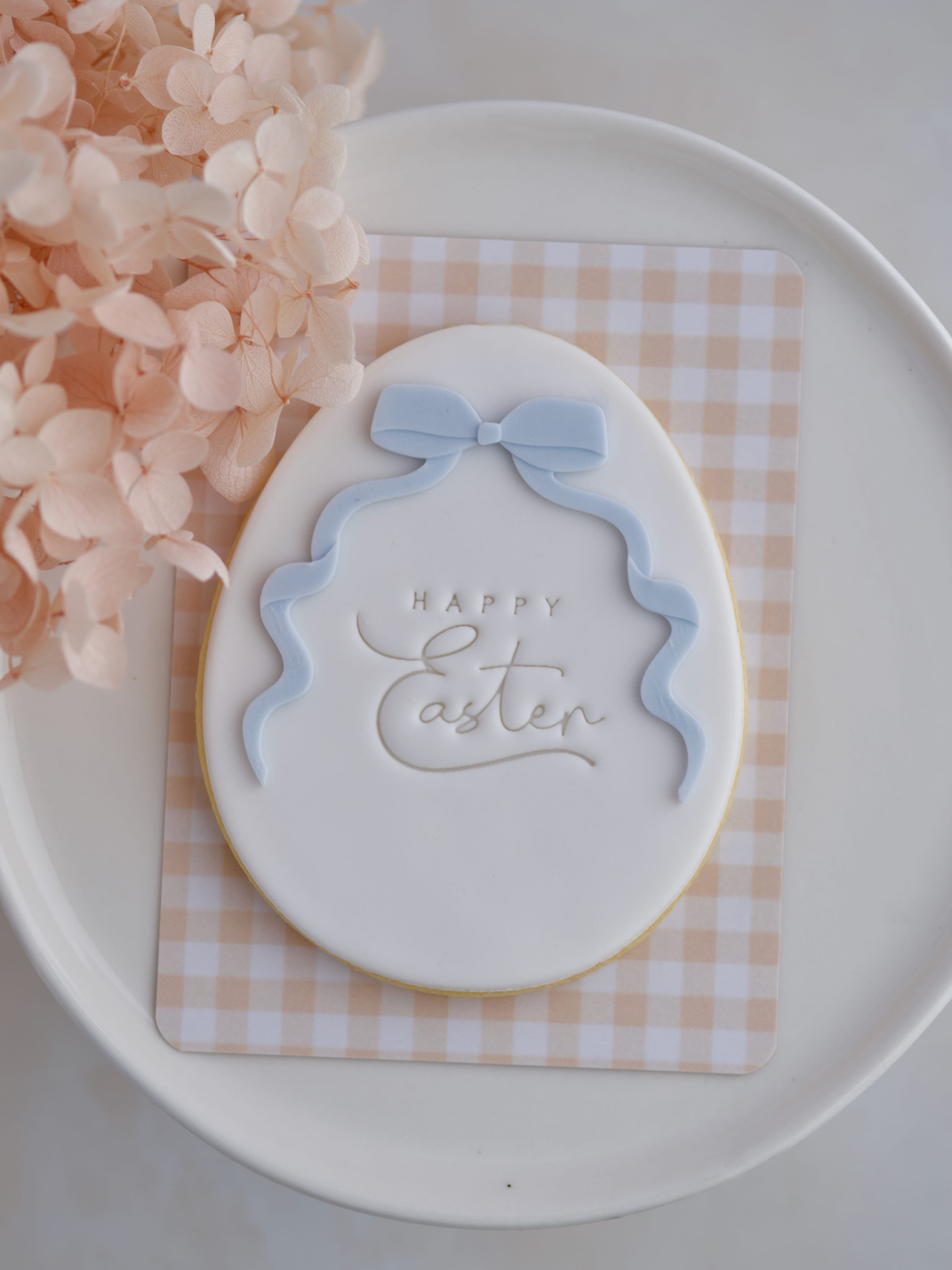 White fondant Easter egg cookie with blue bow and “Happy Easter” stamp.