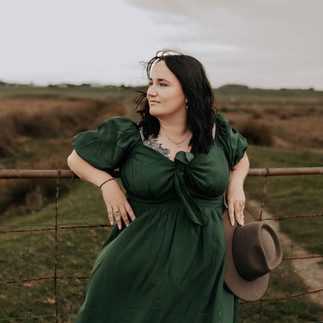Woman in green dress with brown hat in hand leaning on a gate with a dirt track behind her