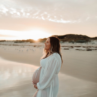 Pregnant woman in white dress holding belly on a beach at sunset