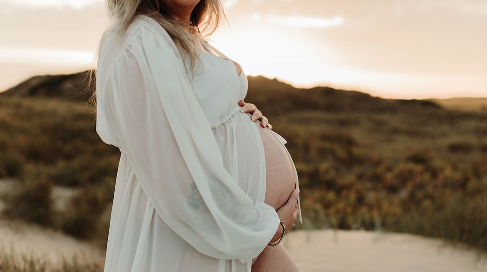 Pregnant woman in a flowy white dress holds her belly in a serene sunset landscape, creating a warm and peaceful mood.