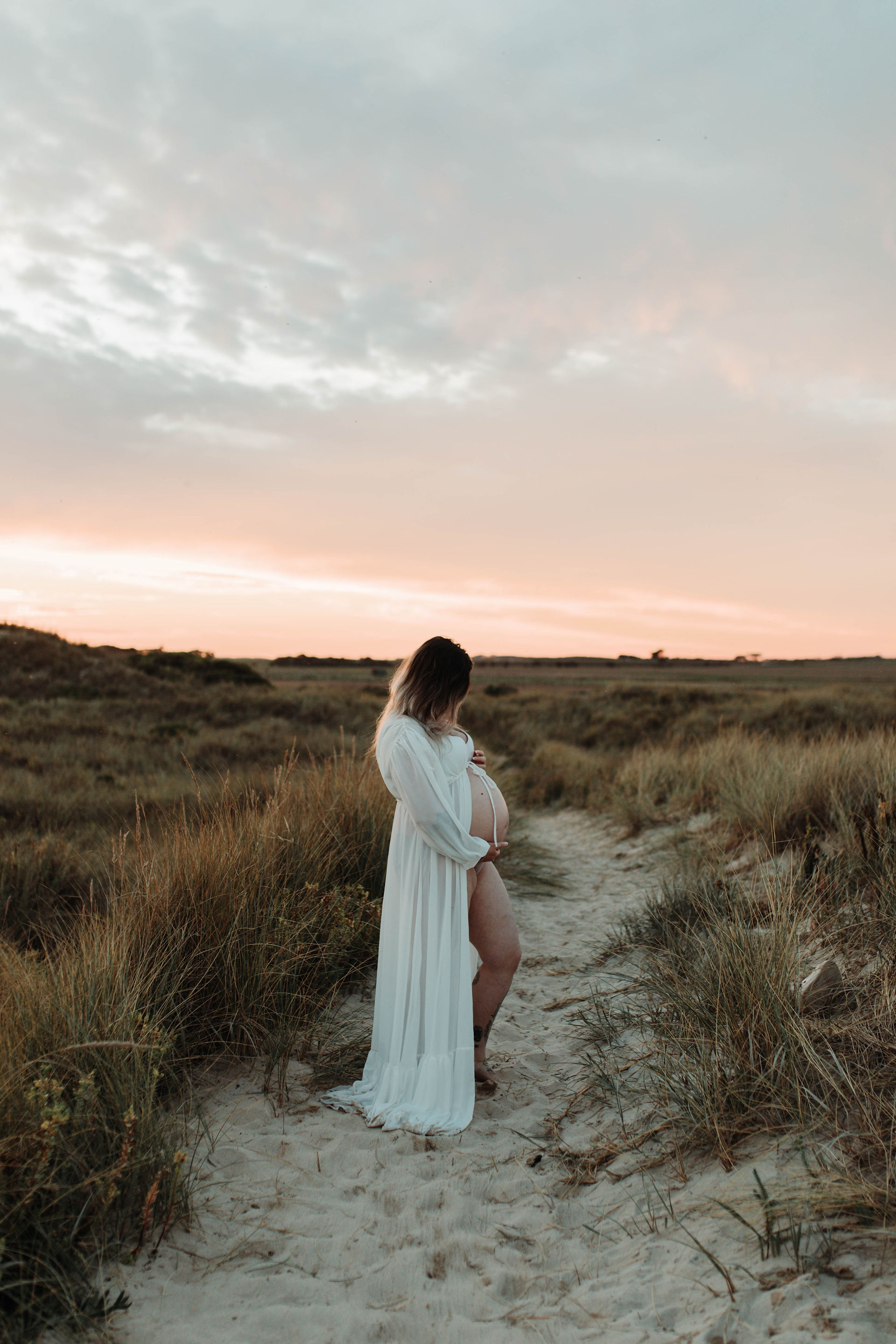 Pregnant woman in white dress holding belly in a grassy landscape