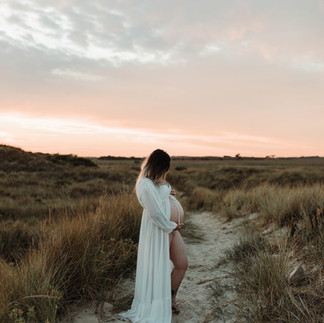 Pregnant woman in white dress holding belly in a grassy landscape