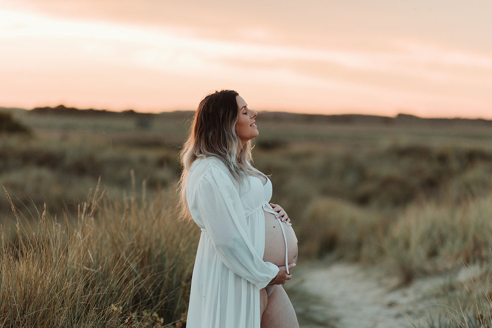 Pregnant woman in white dress stands in a grassy field at sunset, eyes closed, smiling peacefully, with a soft pink sky in the background.