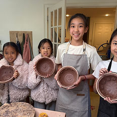 a group of smiling girls wearing aprons, holding clay bowls