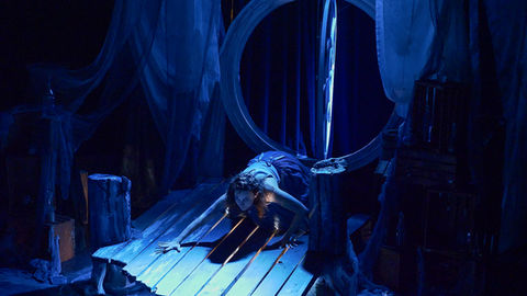 A figure lying on a wooden platform bathed in deep blue light with a large circular set piece behind them - Second Skin