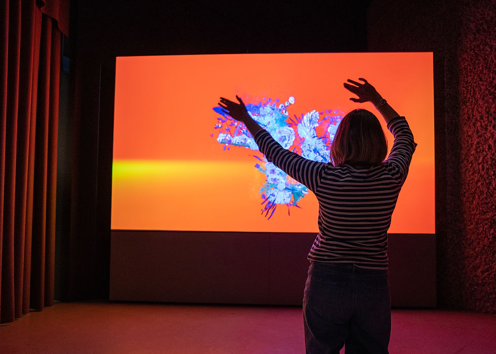 A person in a striped shirt raises their arms toward a bright orange screen with abstract blue patterns in a dark room.