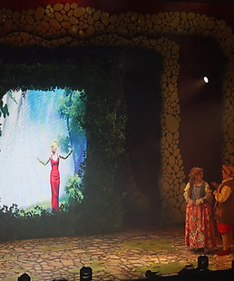 A stage show of a pantomime with a digital screen in the centre featuring a beautiful lady in a red dress