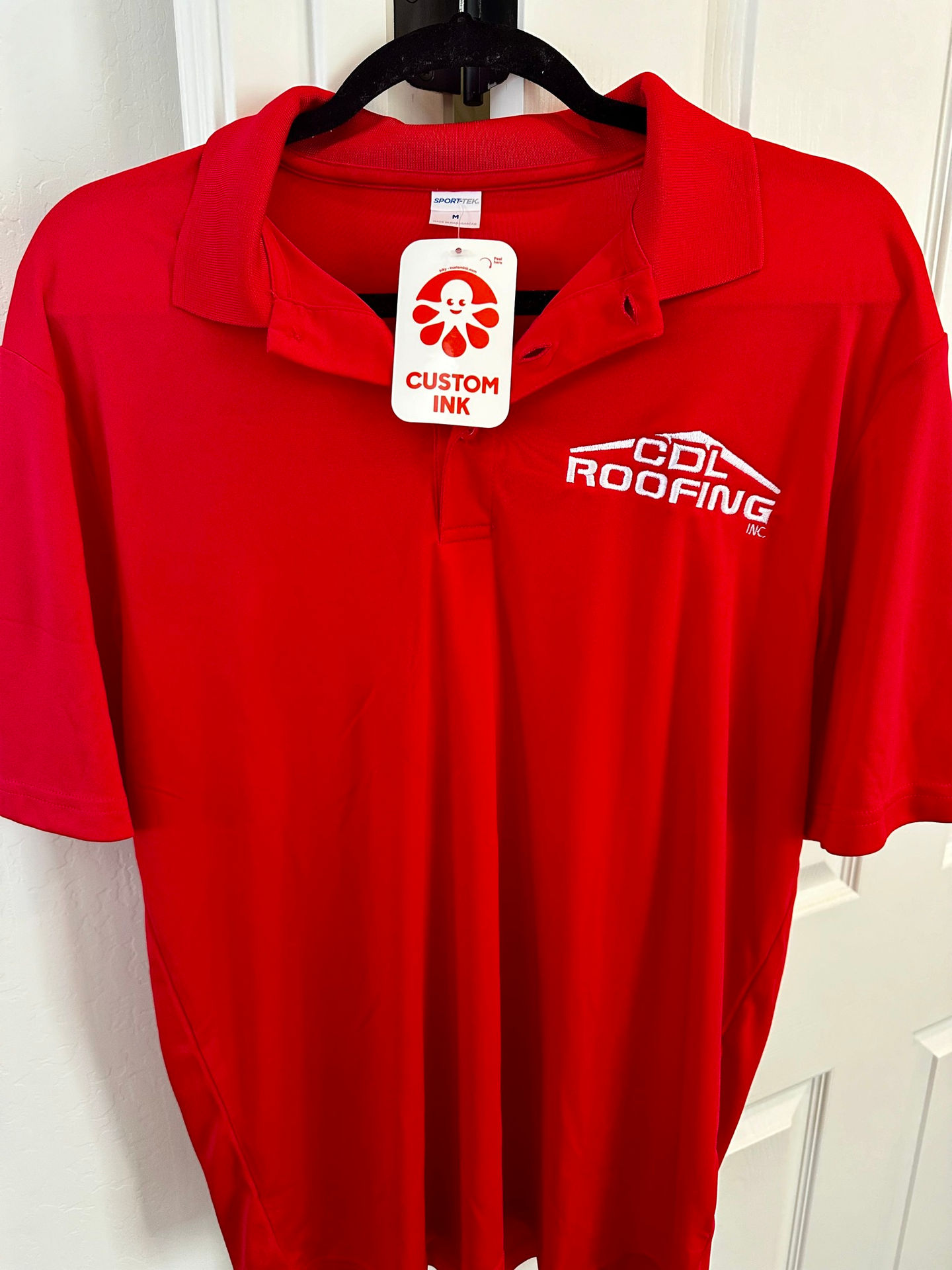 Custom branded polo shirt designed and produced by Sondra Barr for CDL Roofing. The image shows a red Sport-Tek polo shirt with the CDL Roofing logo embroidered on the chest, hanging on a hanger with a Custom Ink tag attached. This represents Sondra Barr’s work in managing custom apparel design and production as part of CDL Roofing’s branding and marketing efforts.