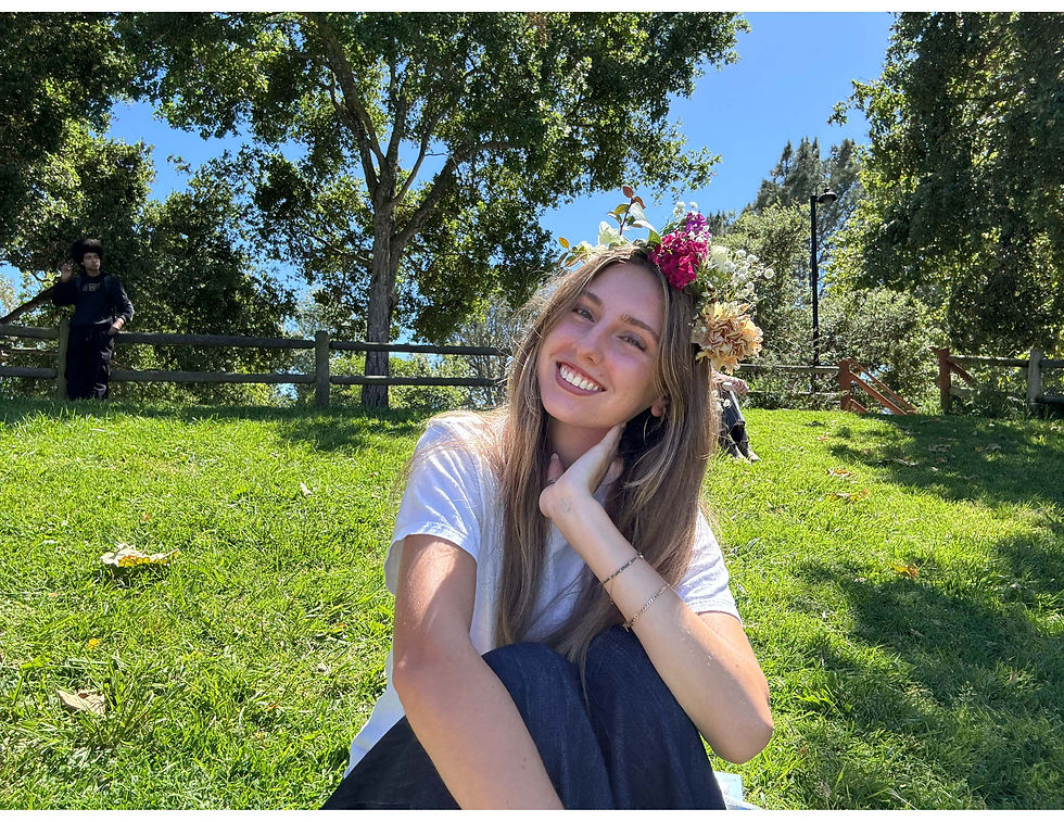 Smiling woman with a flower crown sits on grass in a sunny park, trees in background. Another person walks in the distance. Vibrant and cheerful.