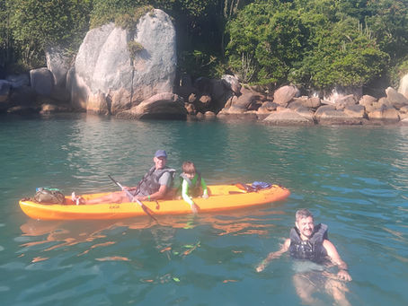 Avô e neto de 5 anos em um caiaque laranja modelo Raia com o pai nadando ao lado, durante passeio guiado da Adventure in Floripa em Canasvieiras, Florianópolis.