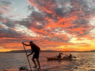 Silhueta de instrutor em pé no Stand Up Paddle e clientes em caiaque duplo durante um nascer do sol com nuvens alaranjadas e avermelhadas no mar de Florianópolis, próximo à Ilha do Francês.