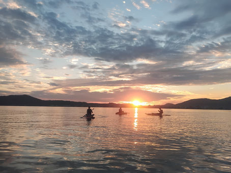 Três pessoas praticando Stand Up Paddle e caiaque em águas calmas durante o nascer do sol. O sol está baixo no horizonte, criando um rastro de luz dourada na água. O céu está decorado com nuvens leves em tons de azul e pêssego, com montanhas ao fundo, transmitindo uma sensação de serenidade e imensidão.