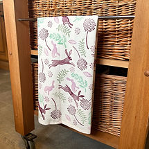 Tea towel printed with block printing stamps in hare and seed head designs in a purple and green colour scheme