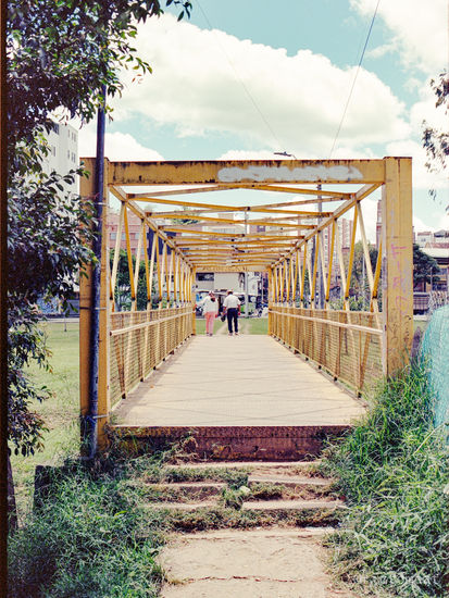 Analog photo of the entrance to a yellow metal bridge in Marinilla, Colombia, with two people walking under a bright sky.
