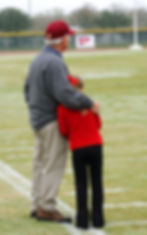 Grandfather hugging his granddaughter on ball field as they watch a game.