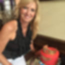 Tina Brickhouse smiling near a red cake with a Wonder Woman theme. She wears a black polka-dot top. The setting seems like a casual indoor space.