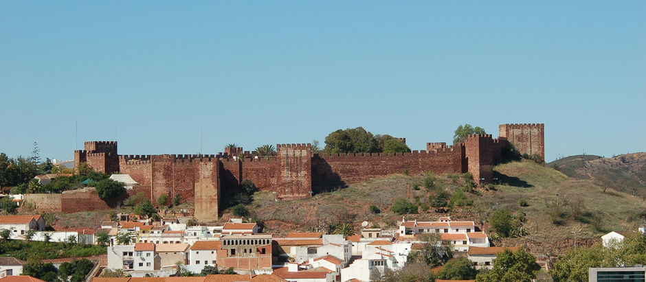 Silves' red-brick castle on a hill, overlooking a town with white buildings and orange roofs. Clear blue sky, lush greenery around. Peaceful scene.
