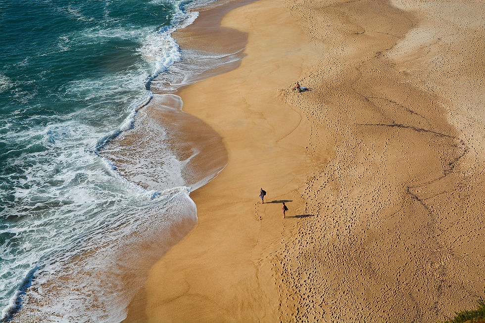 Aerial view of an Algarve sandy beach with two people walking along the shore. Waves crash onto the beach, creating a serene, sunny atmosphere.