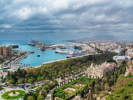 Malagar harbour overlooks a vibrant coastal cityscape with ships in a port and lush gardens. Overcast sky creates a calm, expansive mood.