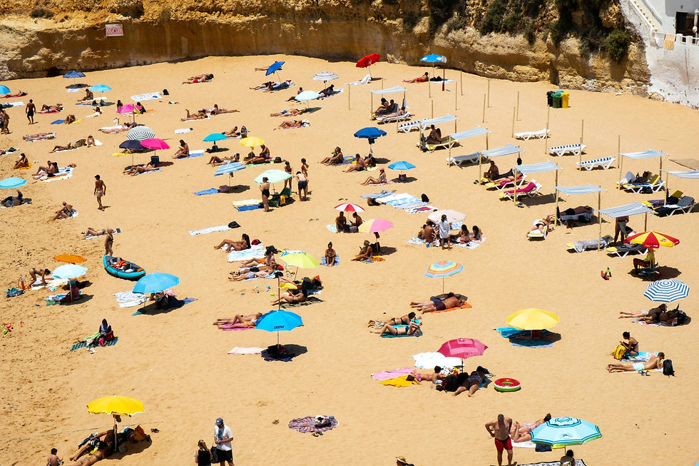 Crowded beach scene in Faro, Algarve, Portugal with people sunbathing under colorful umbrellas on sandy shore. Cliffs in background, relaxing summer vibe.