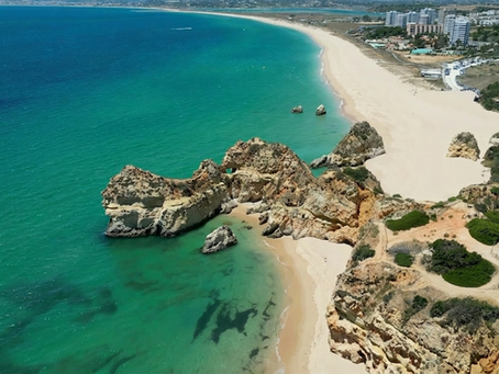 View from Praia dos Três Irmãos looking toward Praia de Alvor, with the Alvor shoreline and estuary stretching into the distance. Turquoise blue water, red rock cliffs and wide, empty sandy beach.