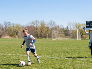 A young boy in a grey and blue soccer uniform dribbles a soccer ball across a bright green field during a sunny daytime game in Ontario, Canada. In the background, a woman wearing a wide-brimmed sun hat and a long-sleeved light grey shirt cheers with her arms raised. A soccer net, a digital scoreboard, and a Canadian flag fluttering on a tall pole are visible behind the players under a clear blue sky.
