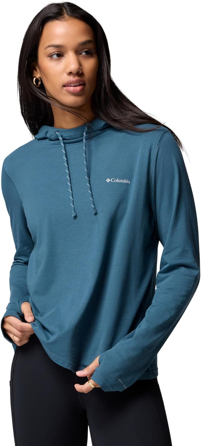 Woman in teal Columbia UPF 50+hoodie holds hem, gazing to the side. Long hair, hoop earrings, black leggings. Calm mood.
