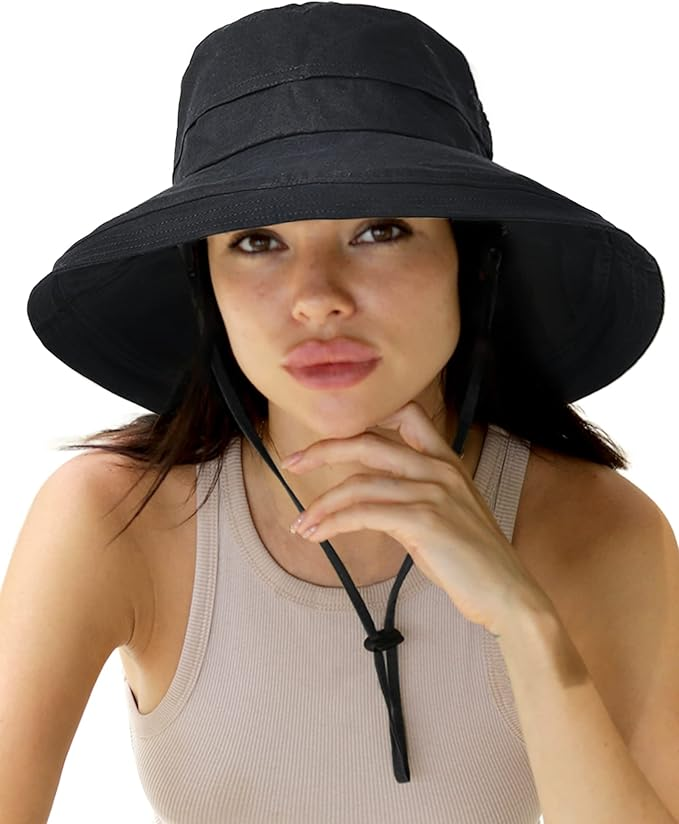 Woman in a tan tank top wearing a Hattis black sunhat with a chinstrap, looking at the camera with a thoughtful expression. White background.