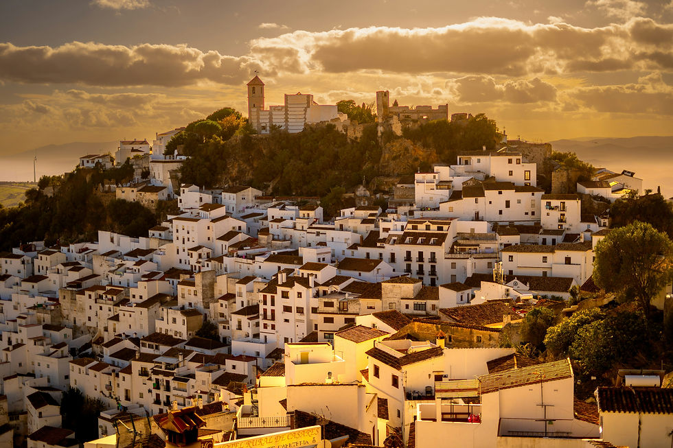 Hilltop village of Casares with white buildings at sunset, surrounded by trees. A tall church tower stands out against a golden sky and clouds.
