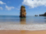 Beach scene at Praia Dona Ana with a tall, rocky sea stack in clear blue water, under a partly cloudy sky. The sandy shoreline shows gentle waves.