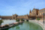 Plaza de España in Seville, Spain, with arched buildings, a canal, and a bridge under a clear blue sky. The Spanish flag is visible.