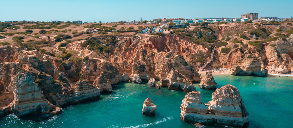 Algarve coast cliffs overlook a turquoise sea with a small boat. A sandy beach with colorful umbrellas lies below. Bright sky enhances the serene mood.