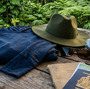 Travel essentials on a wooden table: hat, sunglasses, sunscreen, shirt, umbrella, scarf, and travel documents in a lush green garden setting.
