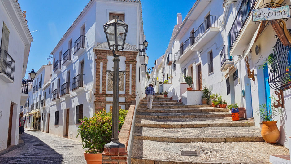 A person walks up stone steps in charming whitewashed village of Frigiliana, Spain with blue sky. Potted plants line the street with a "Locura" sign visible.