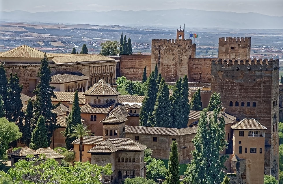 Historic fortress, Alhambra in Granada with stone towers and red roofs surrounded by tall trees. Mountains and countryside in the distant background.