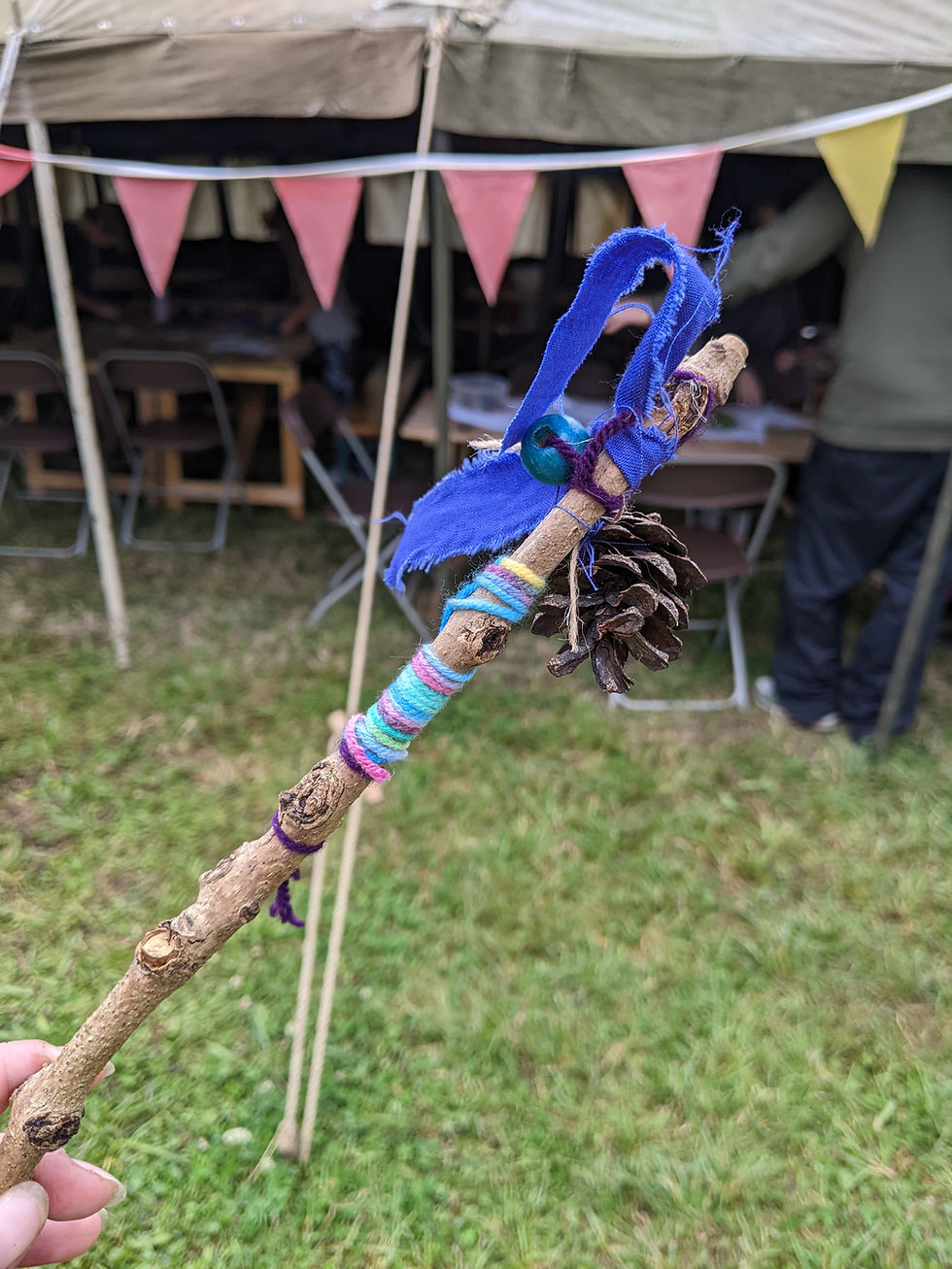 A handmade nature wand made from a stick, decorated with colorful yarn, blue fabric, and a pinecone. It's being held in front of a tent with festive bunting.