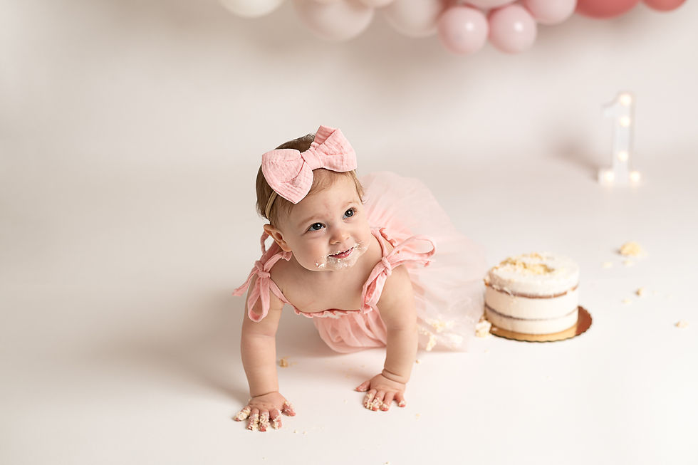 Baby in pink dress crawls on white floor, cake smeared on face and hands. Pink balloons and a lit number "1" in background. Wilmington cake smash photographer