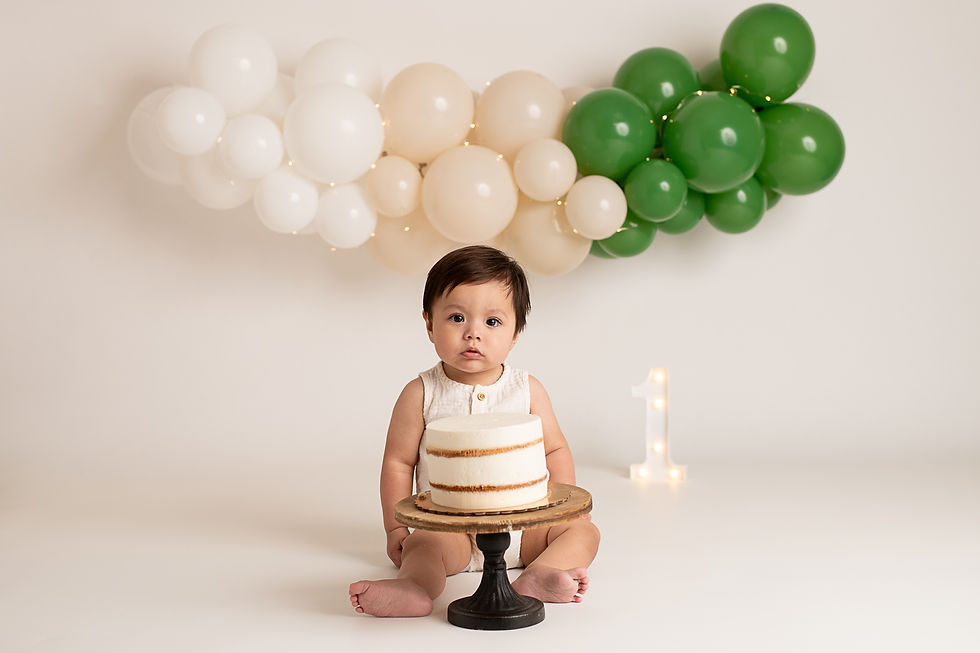 boy seated with a cake, beige and green balloons in background. White "1" on floor. Celebrating first birthday