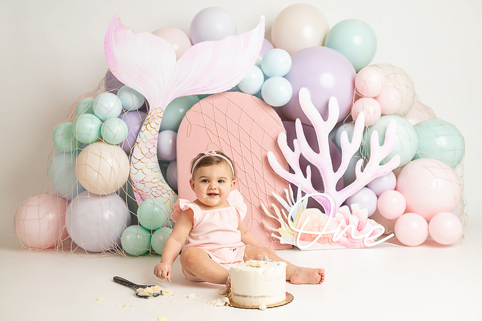 Baby in pink outfit smiles by a cake, surrounded by pastel balloons, a mermaid tail, and under the sea decor. "One" is lit in neon script. Cake smash photographer